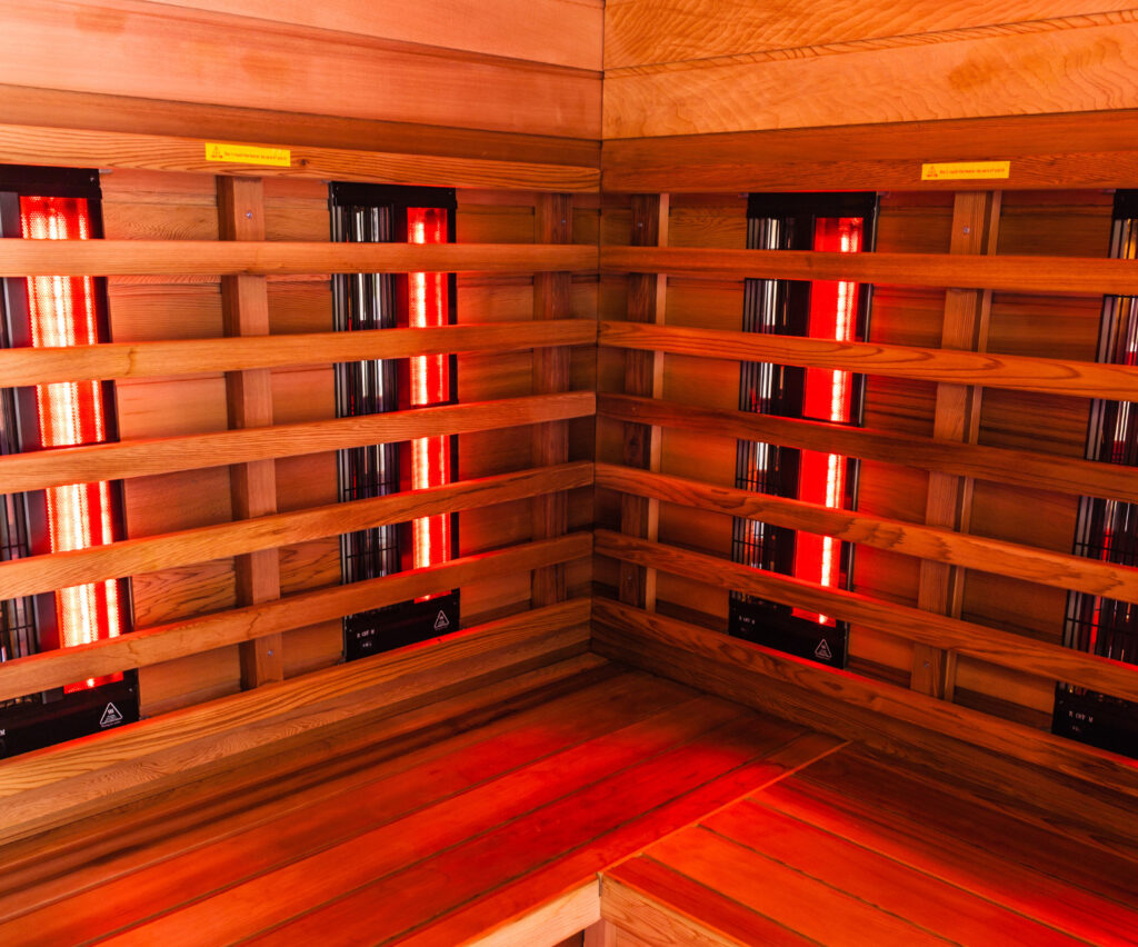 Corner view of a wooden sauna with multiple benches and red infrared heater tubes along the walls.