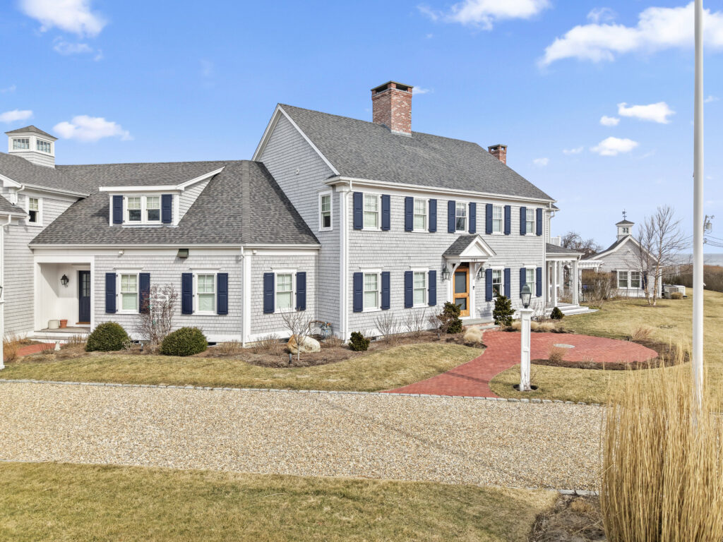 Front exterior drone view of a large white colonial Cape Cod home with dark blue shutters, gray roof shingles, brick entry path, gravel driveway, and early spring landscaping, showcasing a Betterwood Homes addition and renovation project