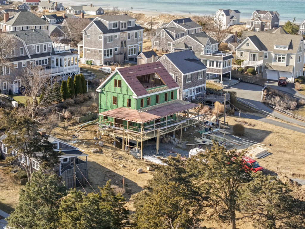 Custom home under construction on Cape Cod with green house wrap, elevated on pilings in a coastal neighborhood